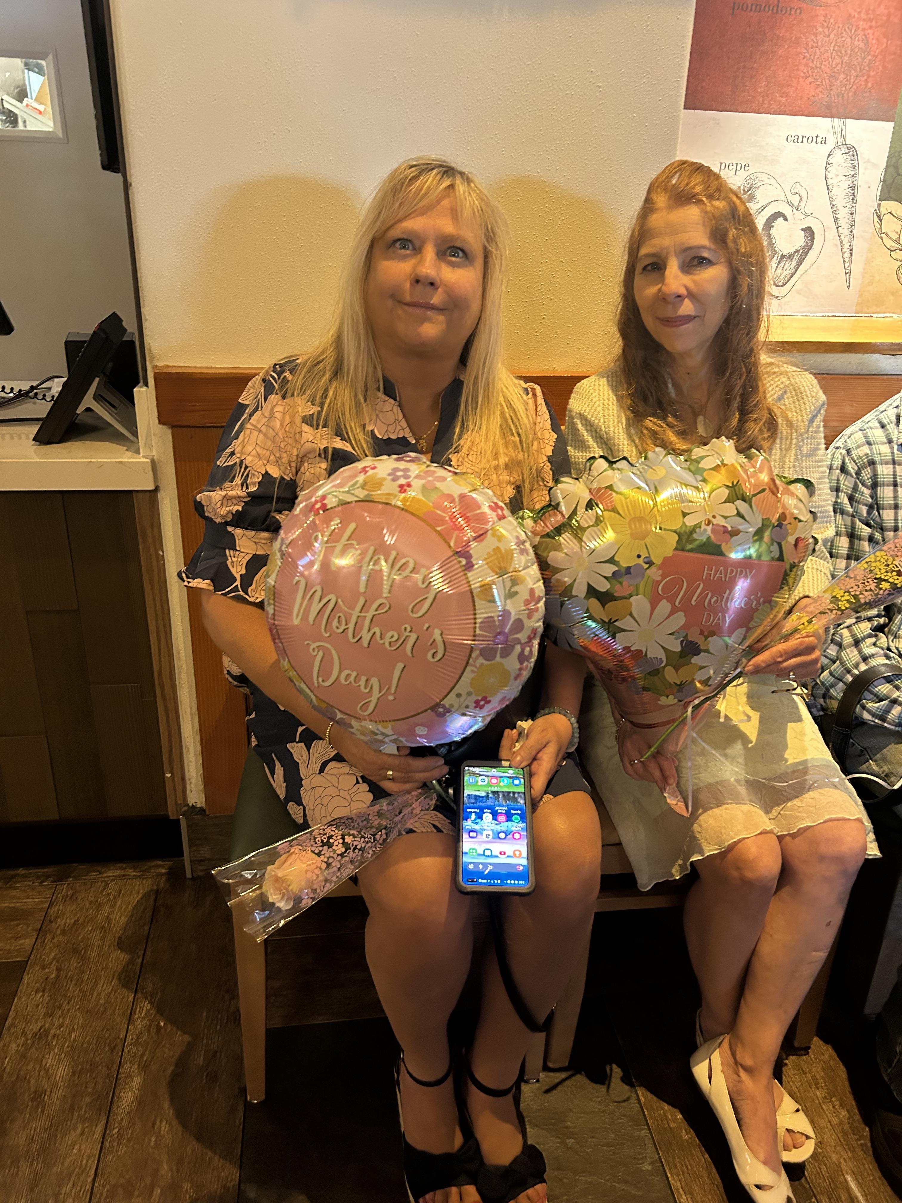 Two women sitting on a bench holding balloons and a cell phone