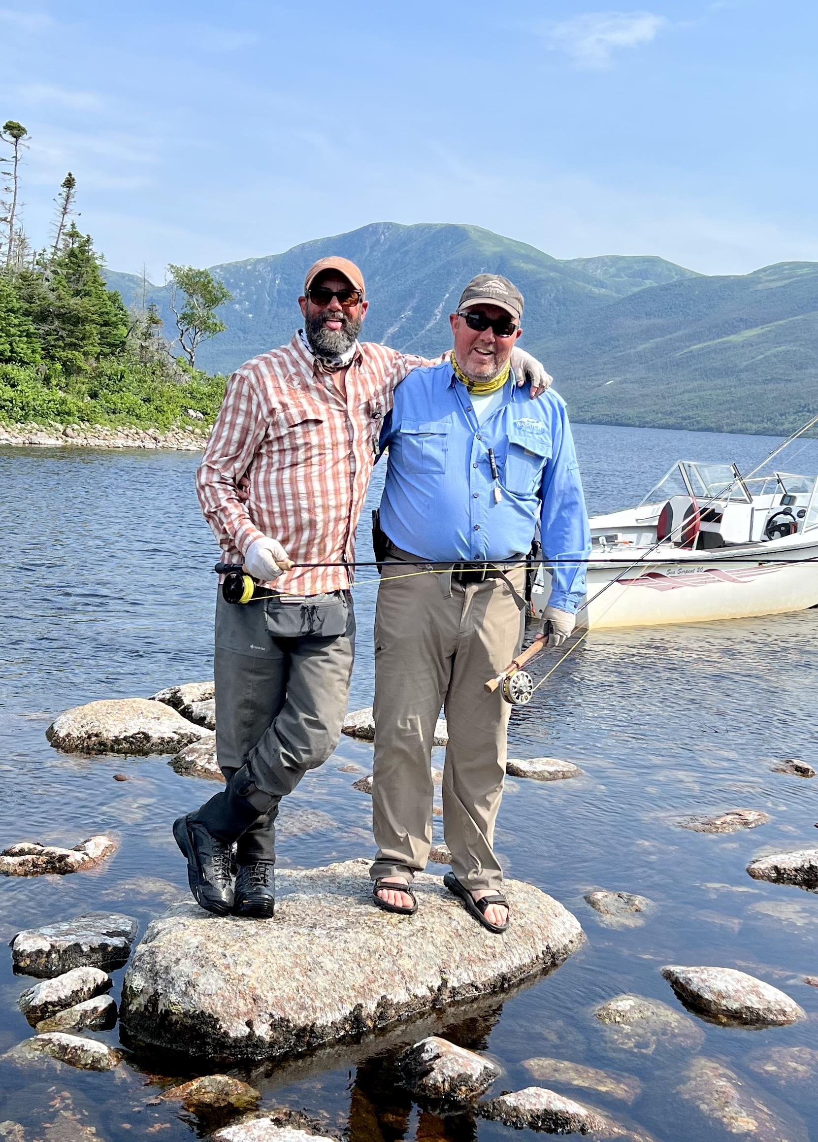 Two men standing on rocks in water with a boat in the background