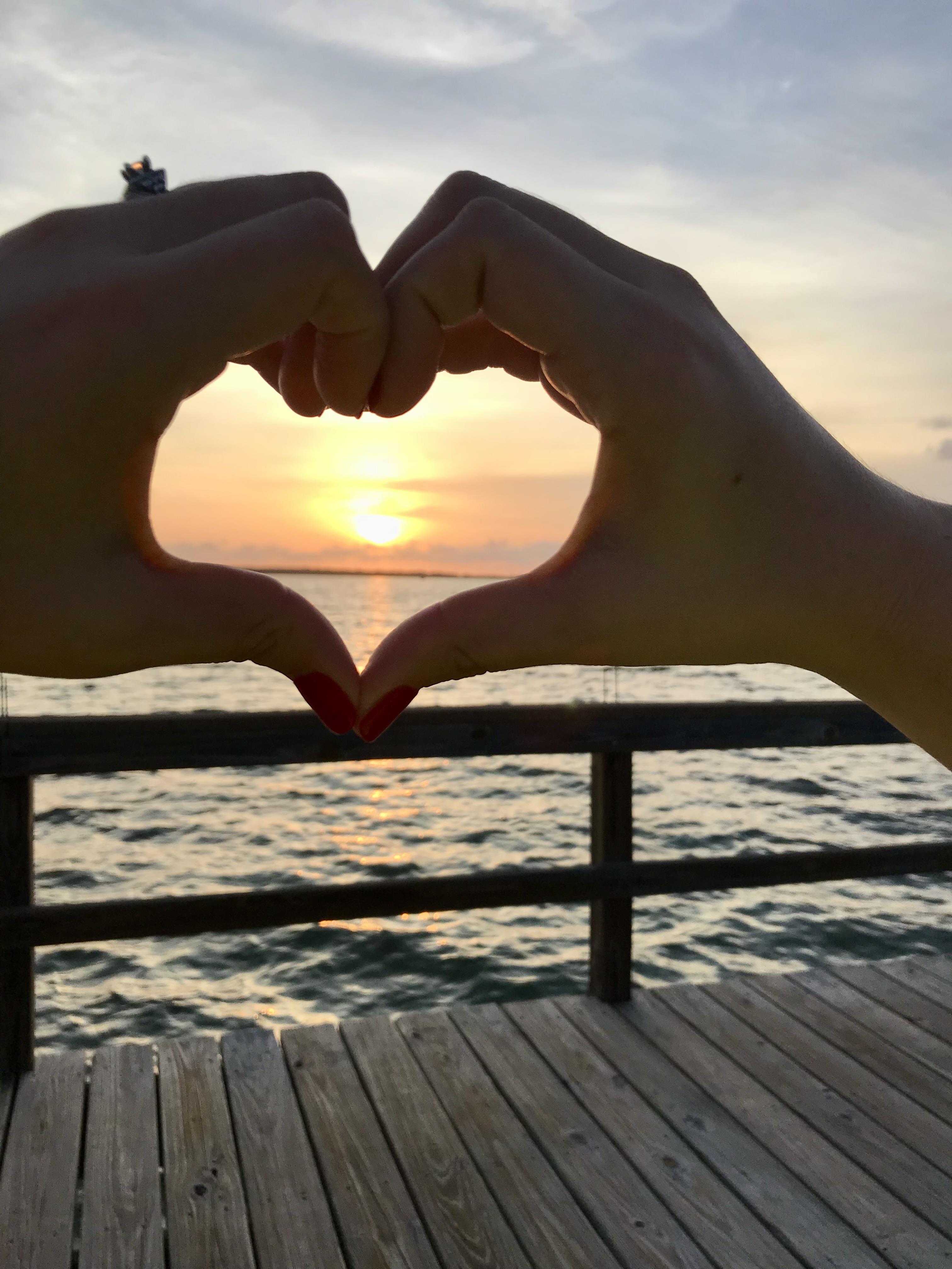 A hand making a heart shape with the sun setting behind it