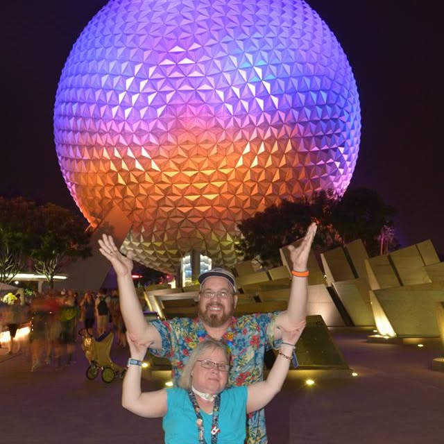 A man and woman posing in front of a large sphere with Epcot in the background