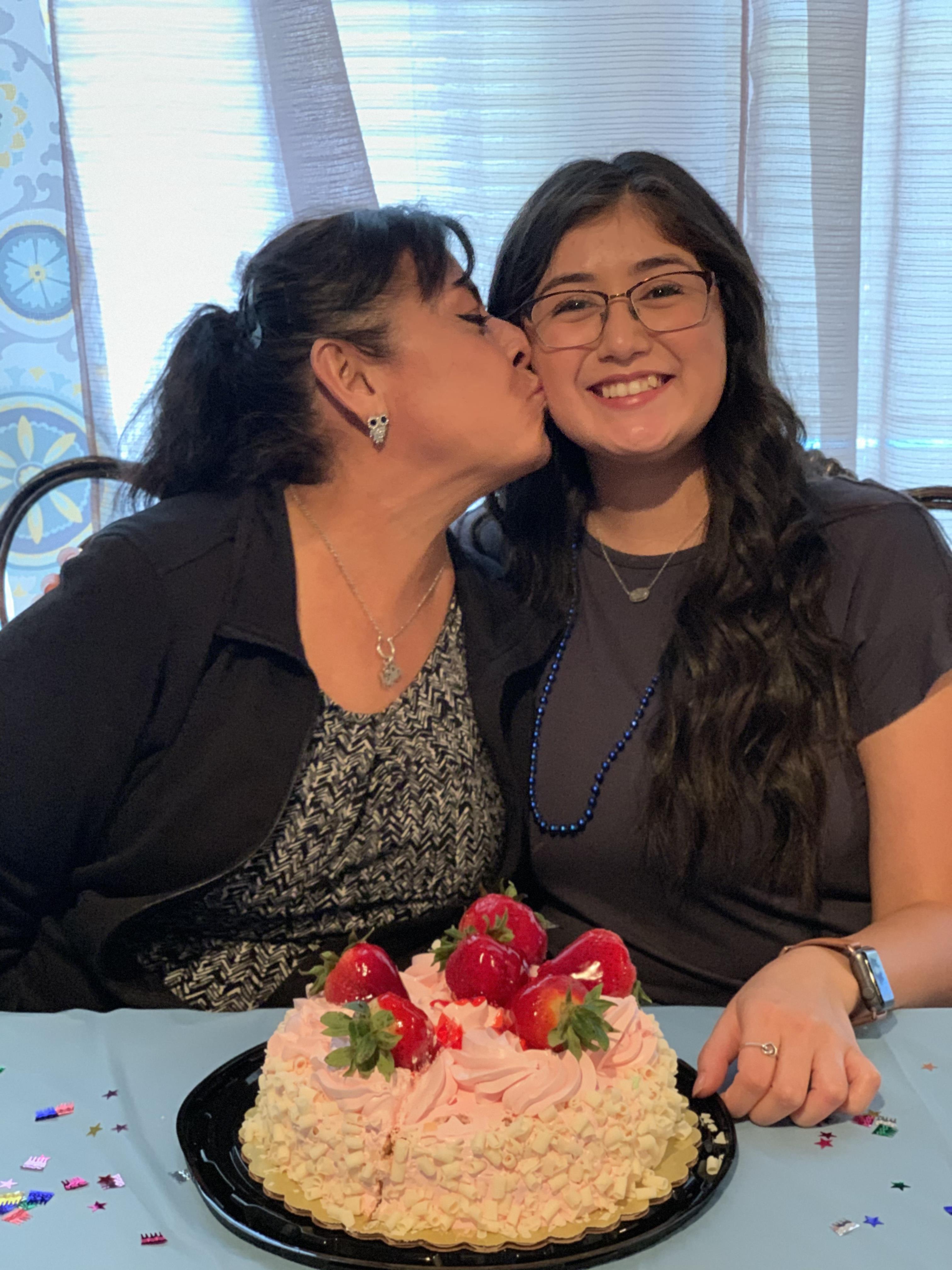 Two women sitting at a table kissing a cake