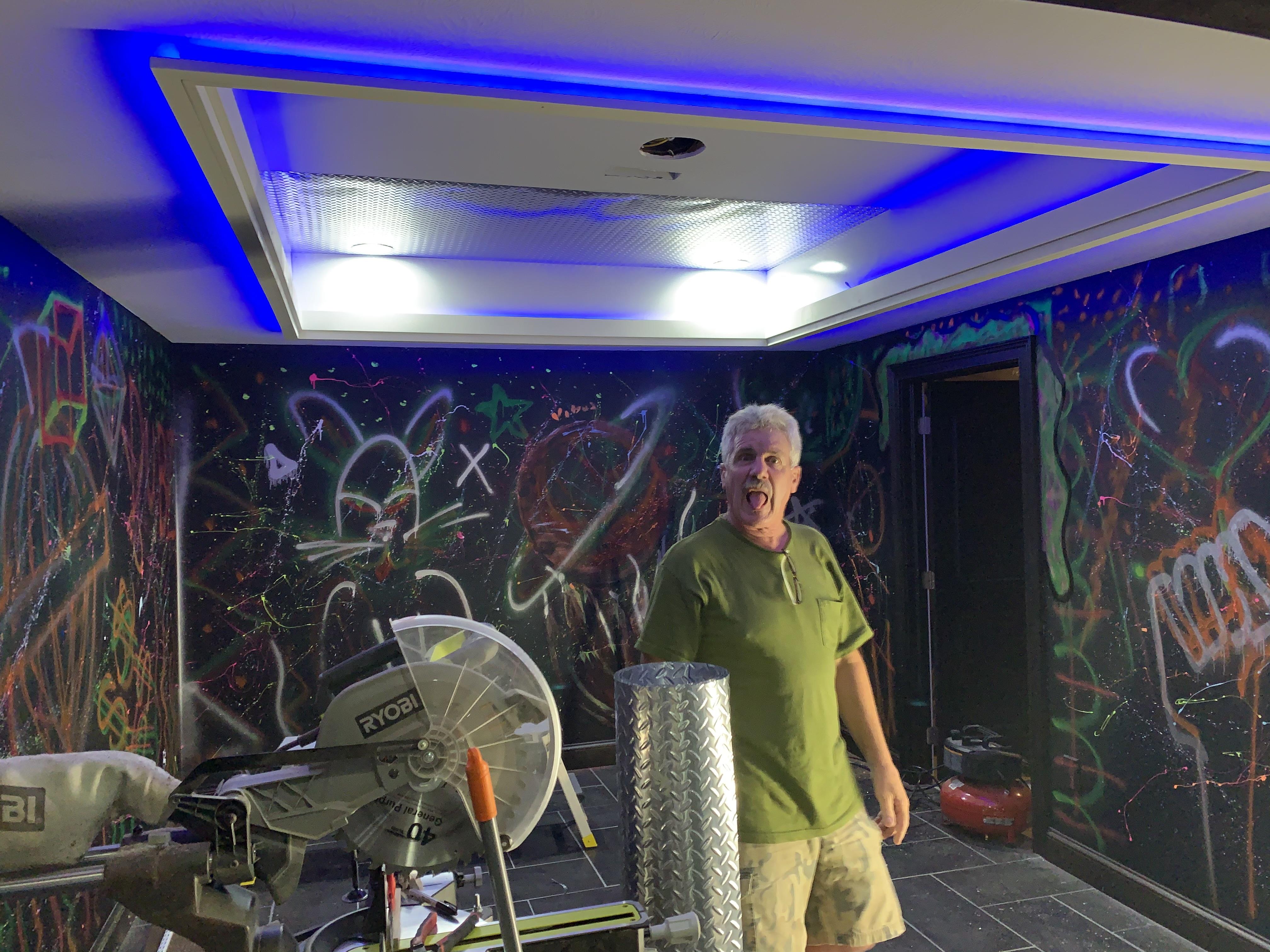 A man standing in a room with a fan and a black wall with graffiti