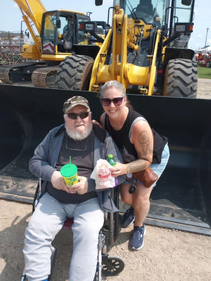 A man and woman sitting in a wheelchair with a bulldozer behind them