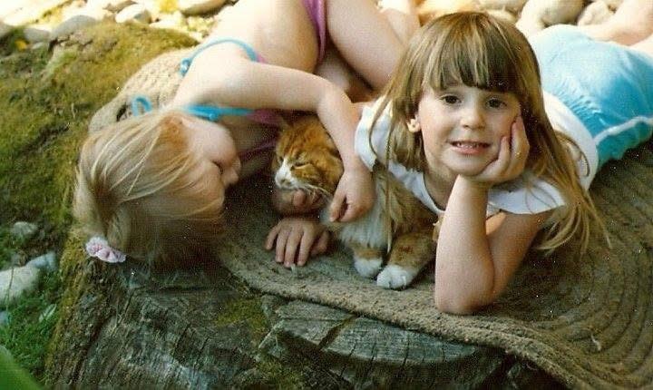 Two young girls cuddle with an orange cat while relaxing on a large tree stump outside.
