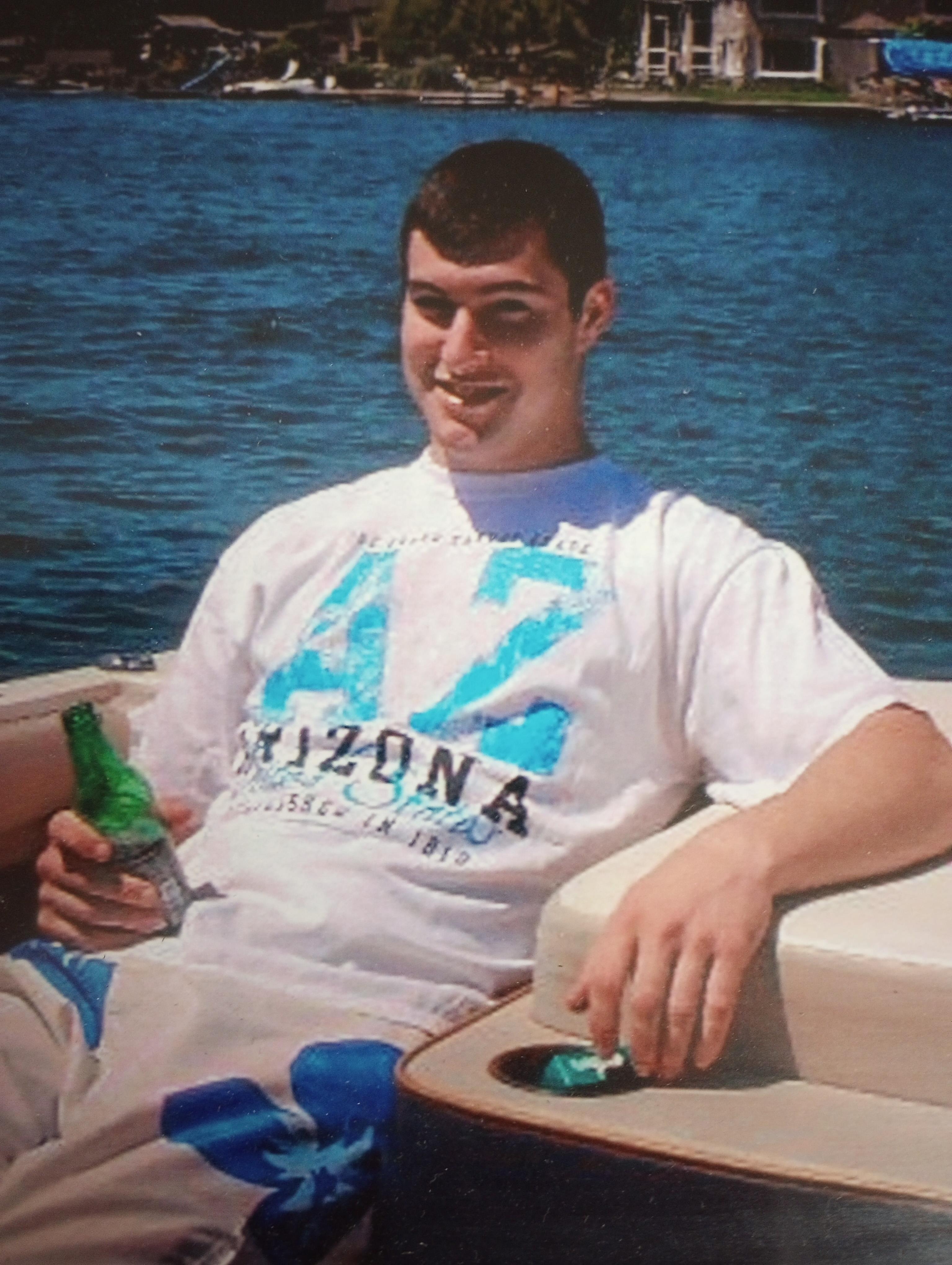 A young man sits on a boat by the lake, holding a green beverage and relaxing in the sun.